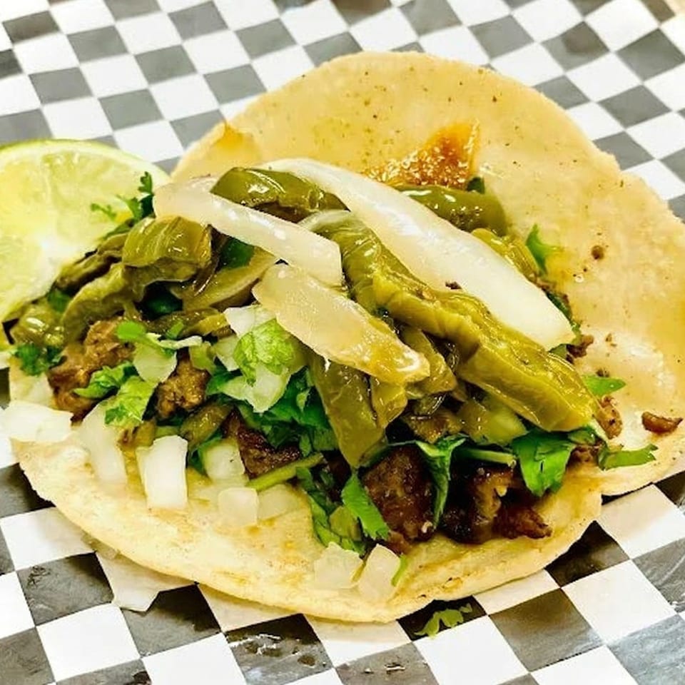 A single taco with meat, green peppers, onions, and cilantro, served with a lime wedge.