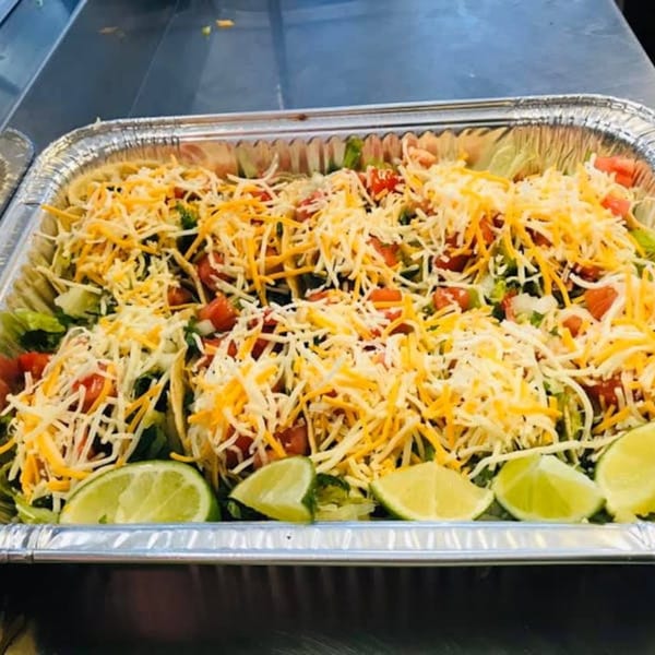 A foil tray filled with tacos topped with shredded cheese, lettuce, tomatoes, and lime wedges.