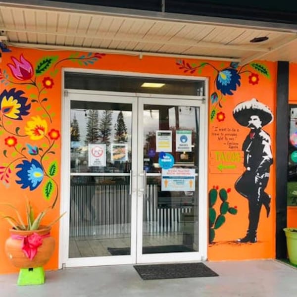 Vibrant orange restaurant exterior with colorful floral murals, glass doors, and a mariachi man painting.