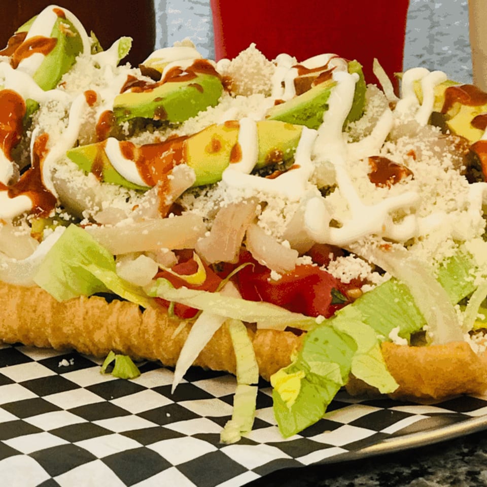 A large, loaded Mexican dish with lettuce, tomatoes, avocado, cheese, and sauces on a checkered paper.