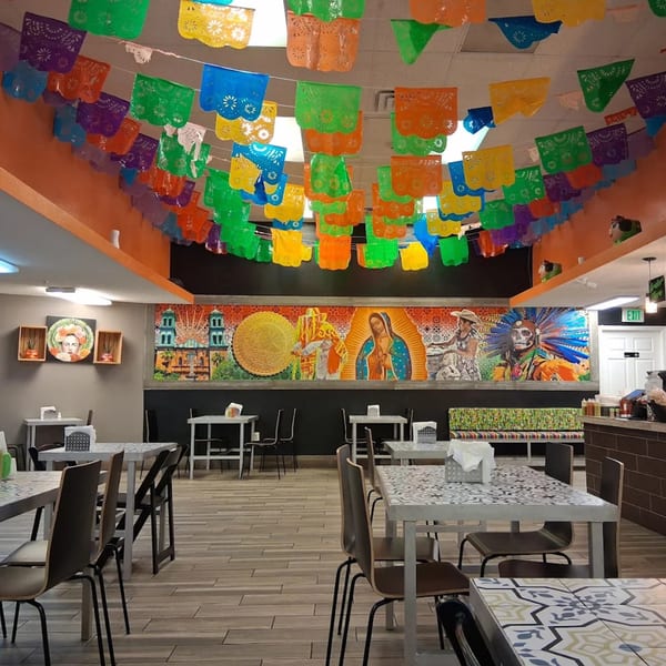 Colorful papel picado banners hang above a vibrant mural and empty tables in a restaurant.