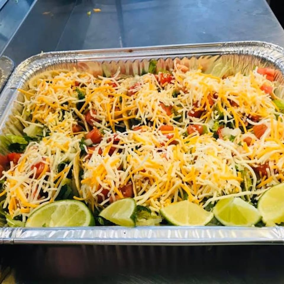 A catering tray filled with tacos topped with shredded cheese, lettuce, tomatoes, and lime wedges.