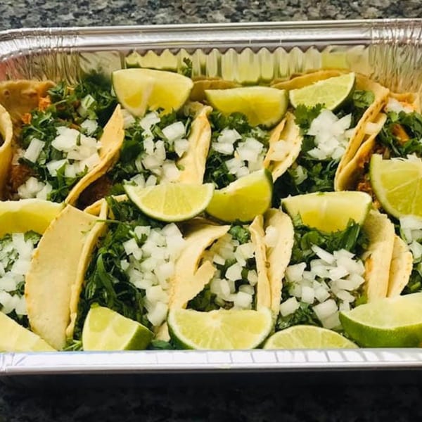 Tacos in a foil tray, topped with cilantro, diced onions, and lime wedges.