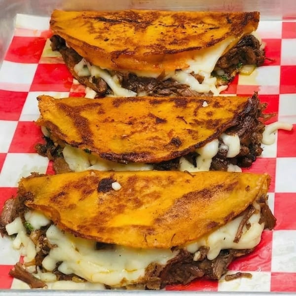 Three cheesy birria tacos with shredded meat on red and white checkered paper.
