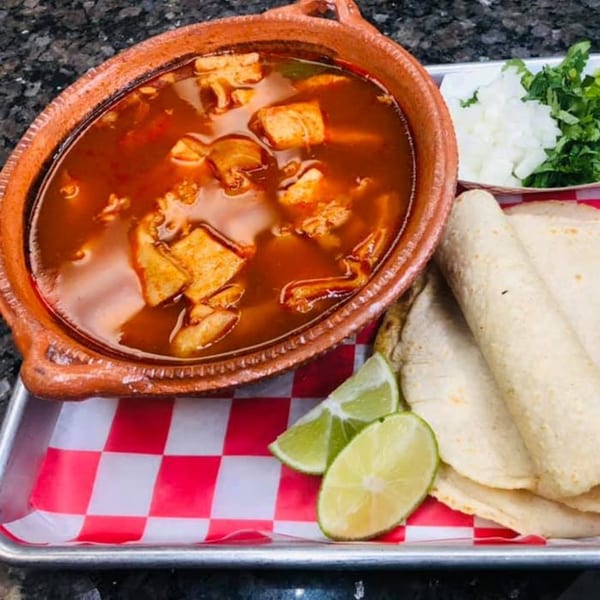 Hearty red stew in a clay bowl with tortillas, lime, diced onions, and cilantro on a tray.